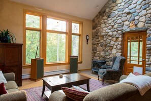 A cozy living room with a stone wall, large windows, and a fireplace. - Unique Nature Inspired Home in Woods next to Lake (Mt Desert)