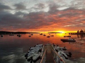 A wooden dock with boats moored at the end, leading to a calm body of water during sunset. - Unique Nature Inspired Home in Woods next to Lake (Mt Desert)