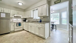 A kitchen with white cabinets, a refrigerator, an oven, and a sink. There is a window with blinds and a door leading to another room. - Just Steps to the Beach! (Virginia Beach)
