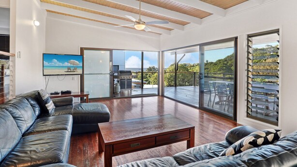 A spacious living area with a leather sofa, a wooden coffee table, and a ceiling fan. Large sliding glass doors lead to an outdoor deck with a view of a lush landscape.