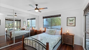A bedroom with a large mirror, a ceiling fan, a bed with floral bedding, bedside tables with lamps, and a window with a view of greenery.