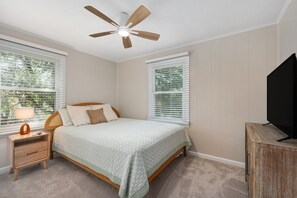 A bedroom with a bed, nightstand, TV stand, ceiling fan, and two windows with blinds. - 364 Close to the Beach and Wright Memorial! (Kill Devil Hills)