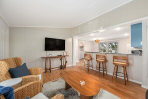A modern living room with a wooden coffee table, wicker sofas, a TV, and a kitchen area with bar stools.