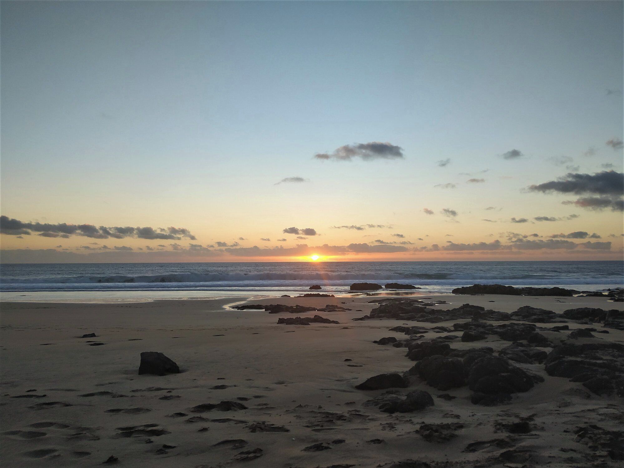 Une plage avec des rochers et du sable, et le soleil se couchant à l’horizon.
