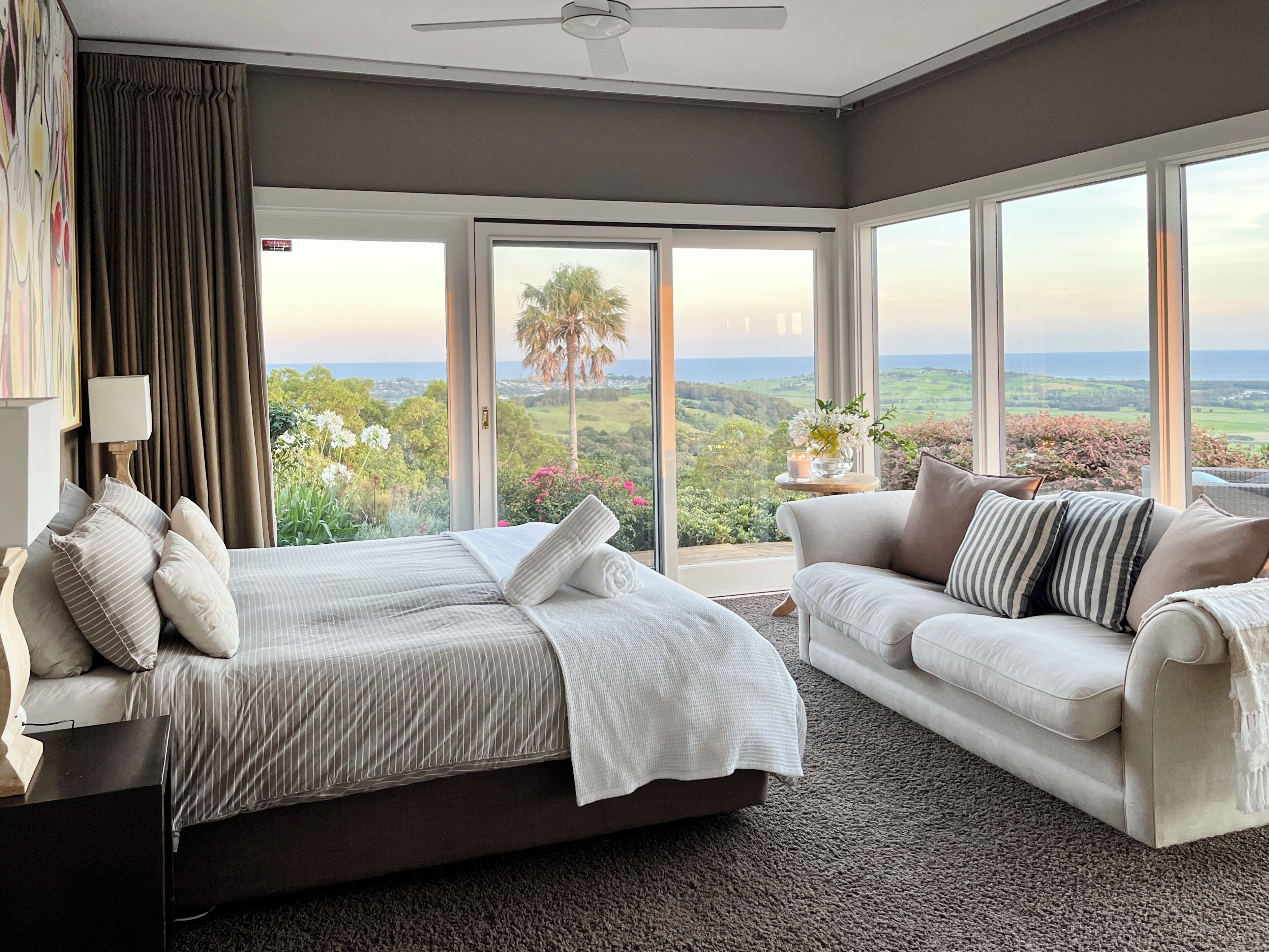 A bedroom with a bed, a sofa, a large window with a view, and a ceiling fan.