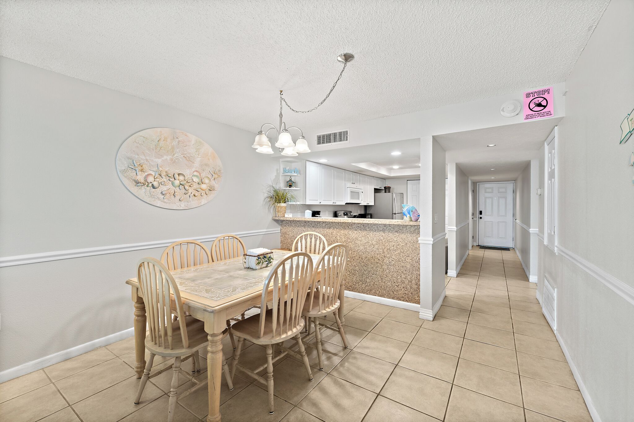 A dining area with a wooden table and chairs, a kitchen in the background, and a hallway leading to a door.