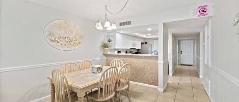 A dining area with a wooden table and chairs, a kitchen in the background, and a hallway leading to a door.