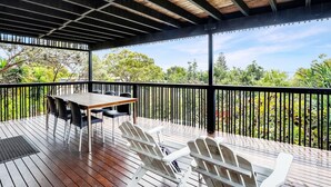 A covered outdoor deck with a table and chairs, surrounded by a black railing, overlooking a lush garden.