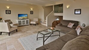A spacious living room with a large sofa, a coffee table, and a television on a cabinet. There is a staircase leading to an upper level.