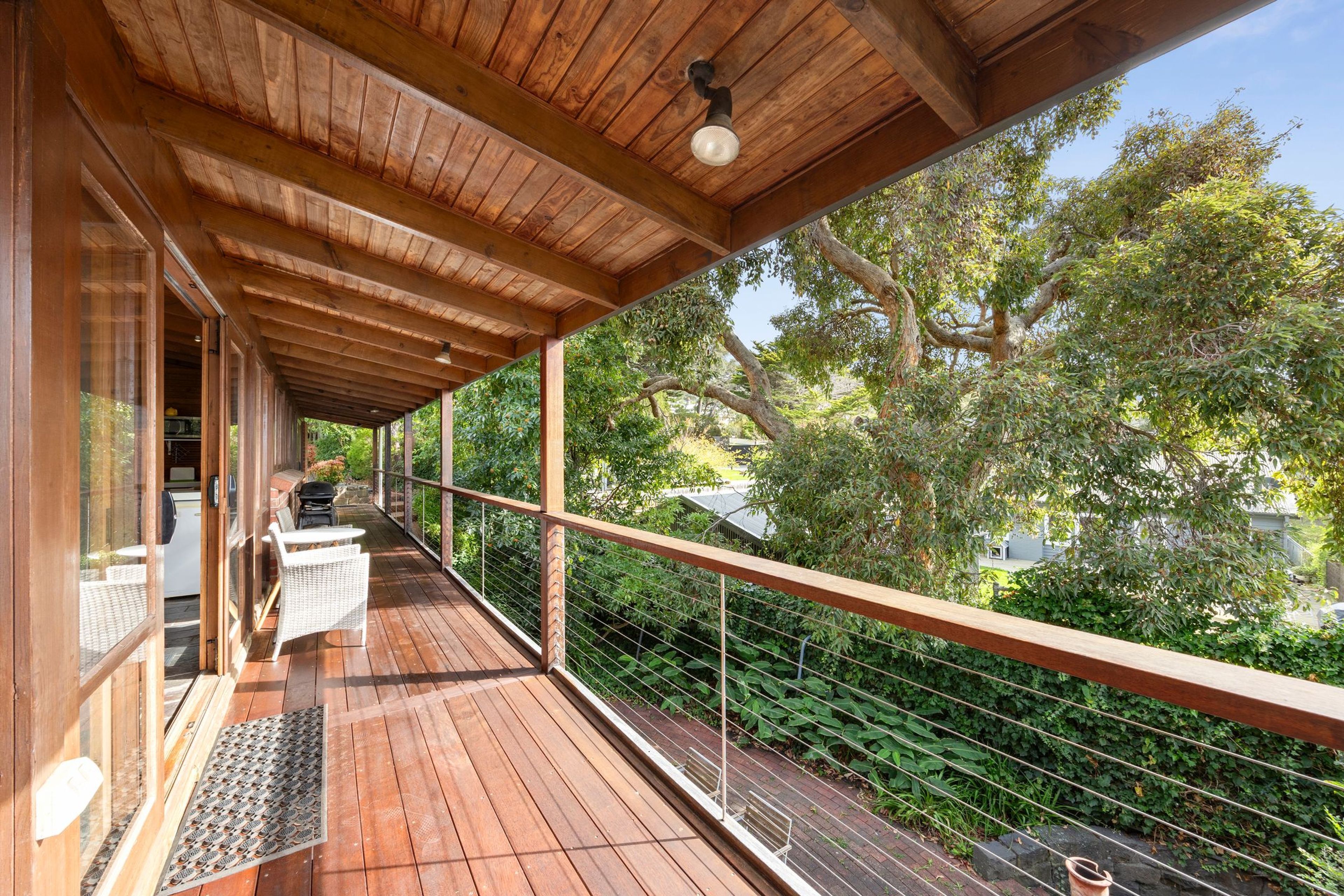 Une terrasse en bois couverte, dotée d’un barbecue et d’un espace salon, qui offre une vue sur un jardin arboré et une maison.