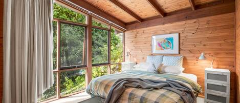 A bedroom with a large window, a bed with a plaid blanket, a nightstand, and a painting on the wall.