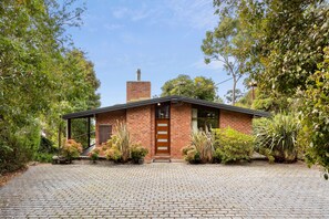 A brick house with a chimney, surrounded by a garden with various plants and a paved driveway. - Bay Vista (Lorne)
