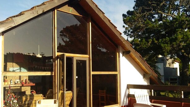 A modern house with a large glass door, a deck with white plastic chairs, and a wooden table. - Hostería Las Grullas (Villa Gesell)