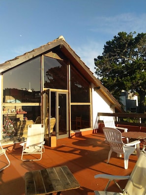 Una casa moderna con un amplio ventanal, una terraza con sillas de plástico blancas y una mesa de madera.