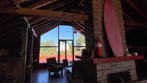 A cozy interior with a brick fireplace, wooden beams, and a view of the outdoors. - Hostería Las Grullas (Villa Gesell)