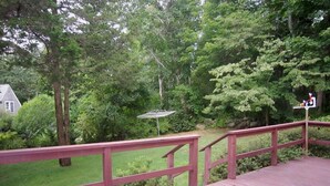 A wooden deck with a red railing overlooking a lush green landscape with trees and a house in the background. - 4BR Cape Style Near Saints Landing and Paines Creek (2086) (Brewster)