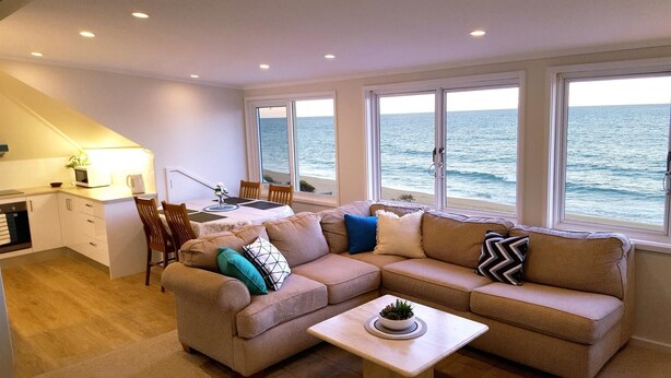 Ein modernes Wohnzimmer mit einem beigefarbenen Sofa, einem Esstisch und einer Küchenzeile. Große Fenster bieten Blick aufs Meer.