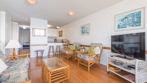 Un salon avec un canapé, des fauteuils, une table basse et un téléviseur. On aperçoit une cuisine au fond.