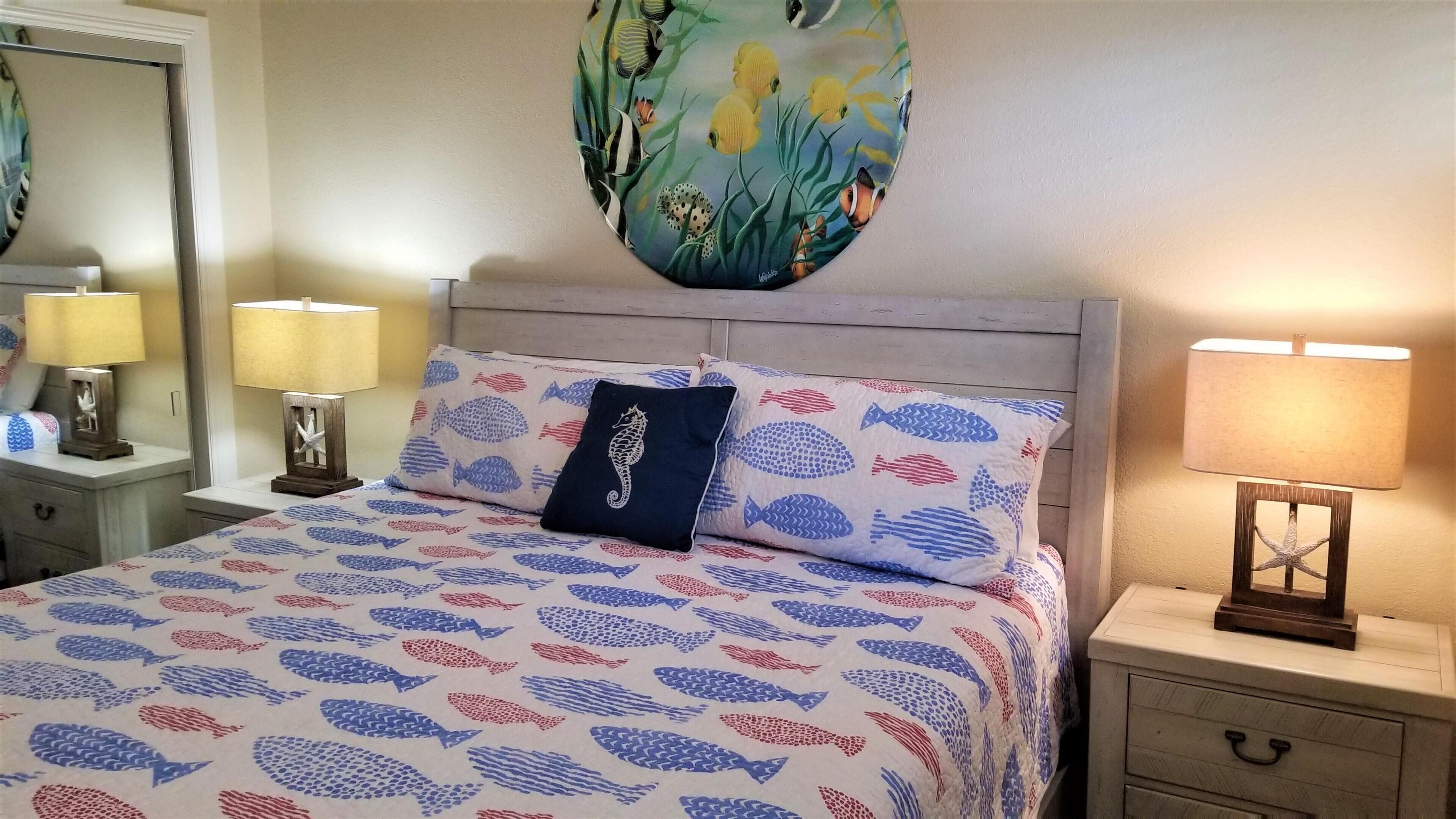 A bedroom with a bed featuring a fish pattern bedspread, two matching pillows, a nightstand with a lamp, and a wall art piece above the bed.