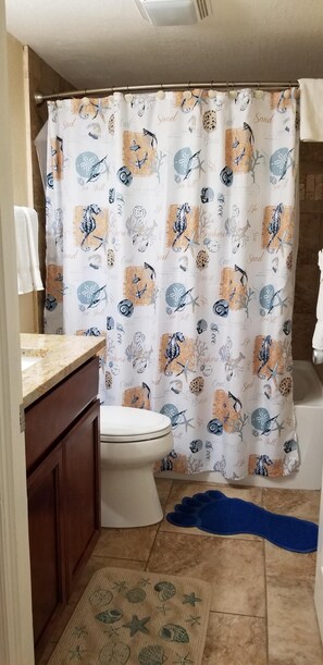A bathroom with a shower curtain featuring sea shell and seahorse patterns, a toilet, a sink with a granite countertop, and a blue bath mat.