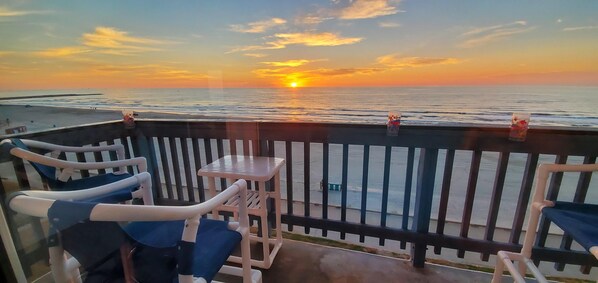 A balcony with a view of the sunset over the sea, equipped with a table and chairs.