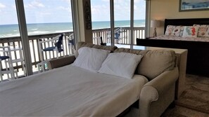 A bed with white linens, a view of the ocean through large windows, and a wooden deck with chairs.