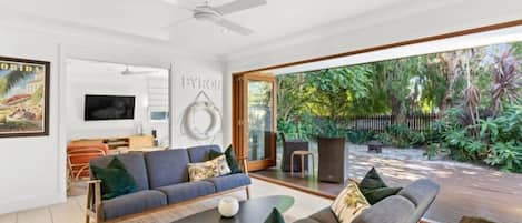 A modern living room with a sofa, coffee table, and a TV. There is a large window with a view of greenery outside.