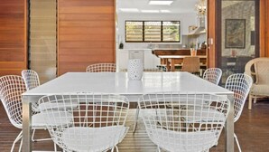 Un coin repas avec une table et des chaises blanches, des murs en bois et une cuisine en arrière-plan.