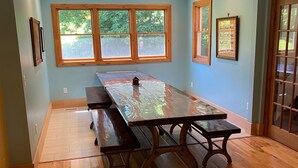 A dining area with a long wooden table, benches, and large windows with a view of greenery.
