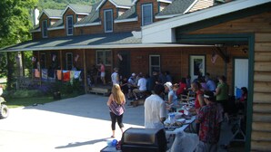 A covered outdoor dining area with tables and chairs, a barbecue grill, and a building with a wooden facade and multiple windows.
