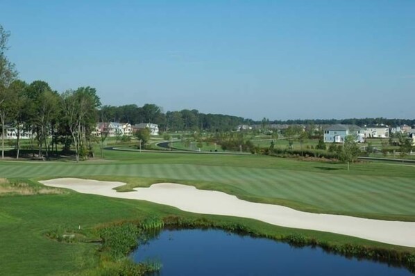Een golfbaan met zandbunkers, een vijver en huizen op de achtergrond.