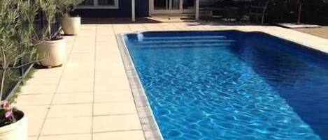 Ein rechteckiger Pool mit gefliestem Deck, ein Haus mit dunklem Dach und Glasfenstern sowie Kübelpflanzen entlang des Decks.