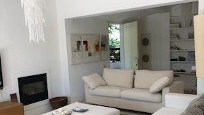 A modern living room with a white sofa, a coffee table, and a chandelier. There is a fireplace and a bookshelf in the background. - Villa Ines, sul mare con piscina (CAPOTERRA)