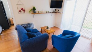 Een woonkamer met een blauwe bank, een blauwe fauteuil, een klein houten tafeltje en een tv aan de muur.
