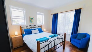 Een slaapkamer met een houten bed, twee nachtkastjes, een blauwe fauteuil en een raam met blauwe gordijnen.