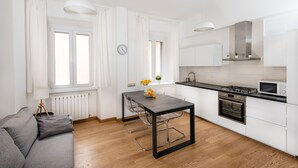 Una cocina moderna con armarios blancos, encimera negra y electrodomésticos de acero inoxidable. Incluye una mesa de comedor con sillas, un sofá y una ventana con cortinas.