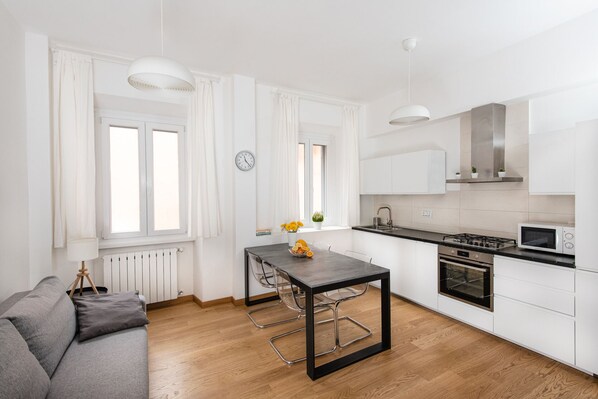 Une cuisine moderne avec des meubles blancs, un plan de travail noir et des appareils en acier inoxydable. On y trouve une table à manger avec des chaises, un canapé et une fenêtre avec des rideaux.