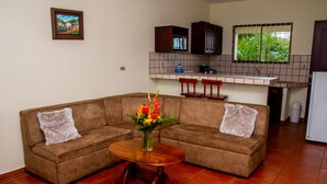 A living room with a brown sectional sofa, a wooden coffee table, and a vase of flowers. There is a kitchen area with cabinets and a window. - Condos Vista Al Volcán (La Fortuna)