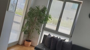 Uma sala de estar moderna com um sofá cinzento, uma mesa pequena branca, uma planta em vaso e janelas amplas com persianas.