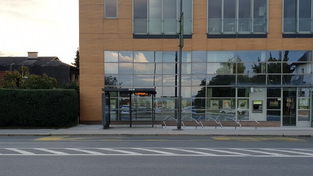 Um edifício moderno com fachada de vidro, um ponto de ônibus e uma rua livre à frente.