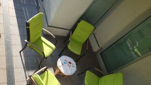Uma pequena área externa com assentos, incluindo quatro cadeiras verdes e uma mesa redonda, tudo em piso de azulejos e com uma janela com persianas.