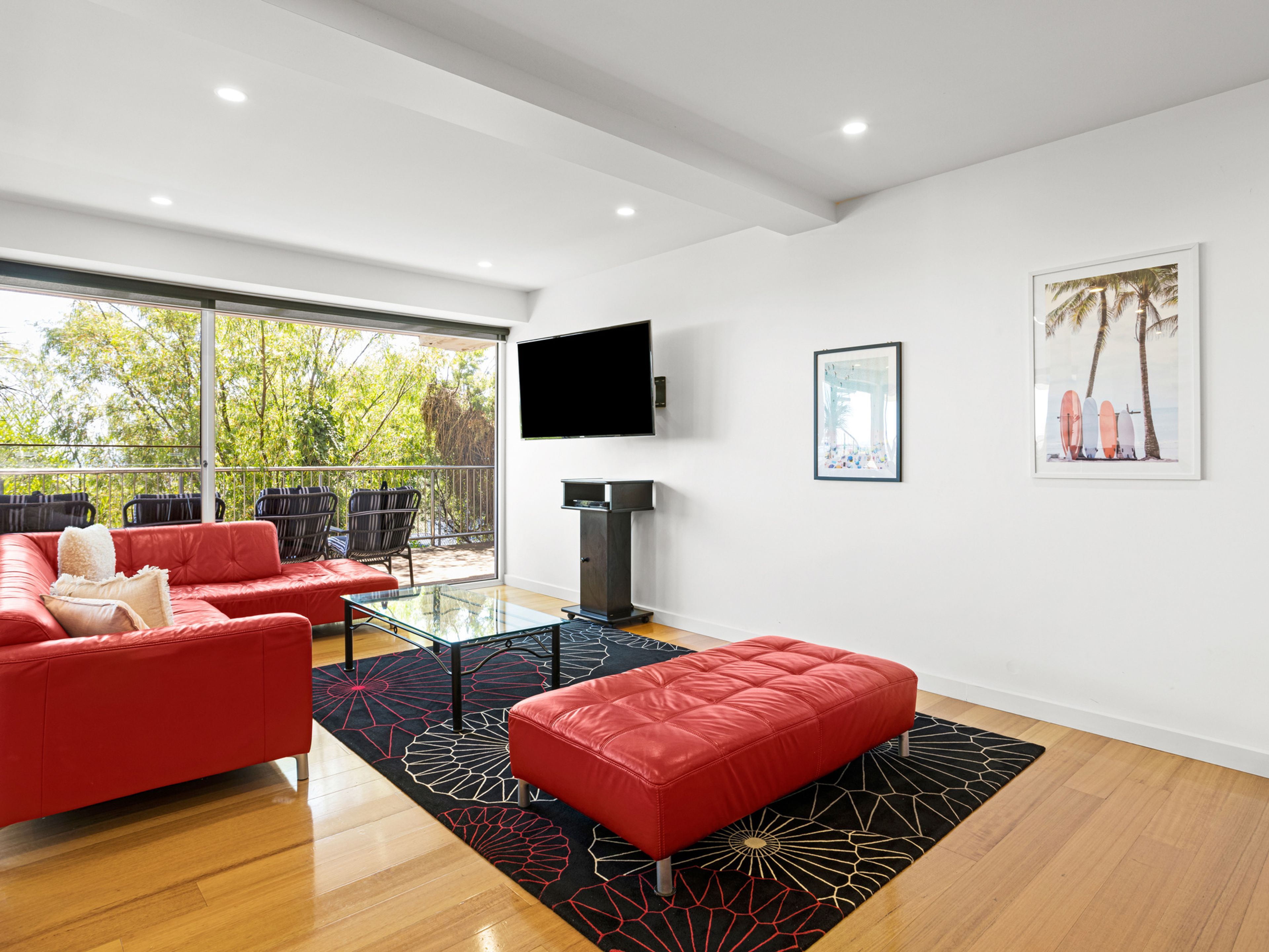 A modern living room with a red sofa, a glass coffee table, and a red ottoman. There is a large window with a view of trees and a balcony with chairs.