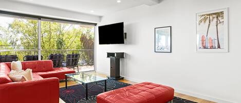 A modern living room with a red sofa, a glass coffee table, and a red ottoman. There is a large window with a view of trees and a balcony with chairs.