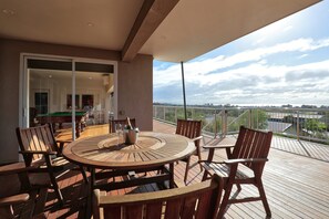 A wooden deck with a round table and chairs, a bottle of champagne, and a view of a river and distant landscape.