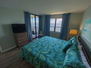 Un dormitorio con cama, televisor, sillón, lámpara y vistas al mar a través de las ventanas.