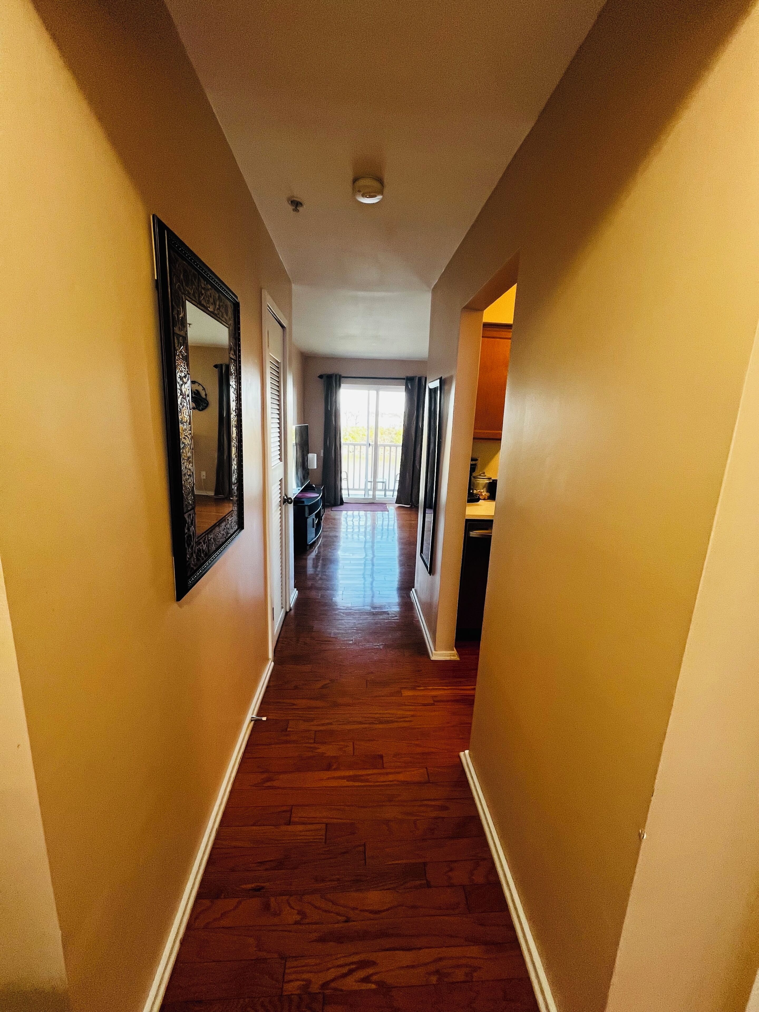 A hallway with wooden flooring, a large mirror on the left, and a door leading to a balcony.