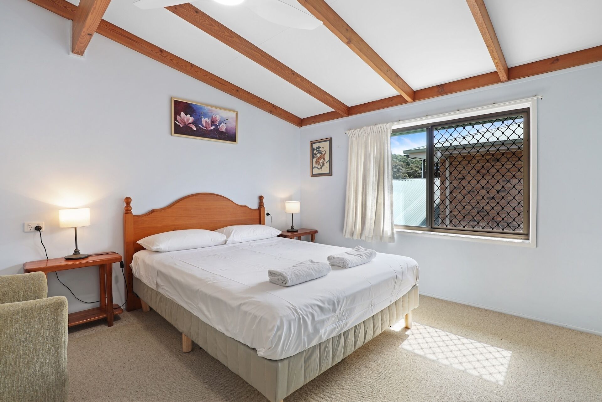 Un dormitorio con una cama de madera, dos mesitas de noche con lámparas, una ventana con cortinas y un cuadro en la pared.