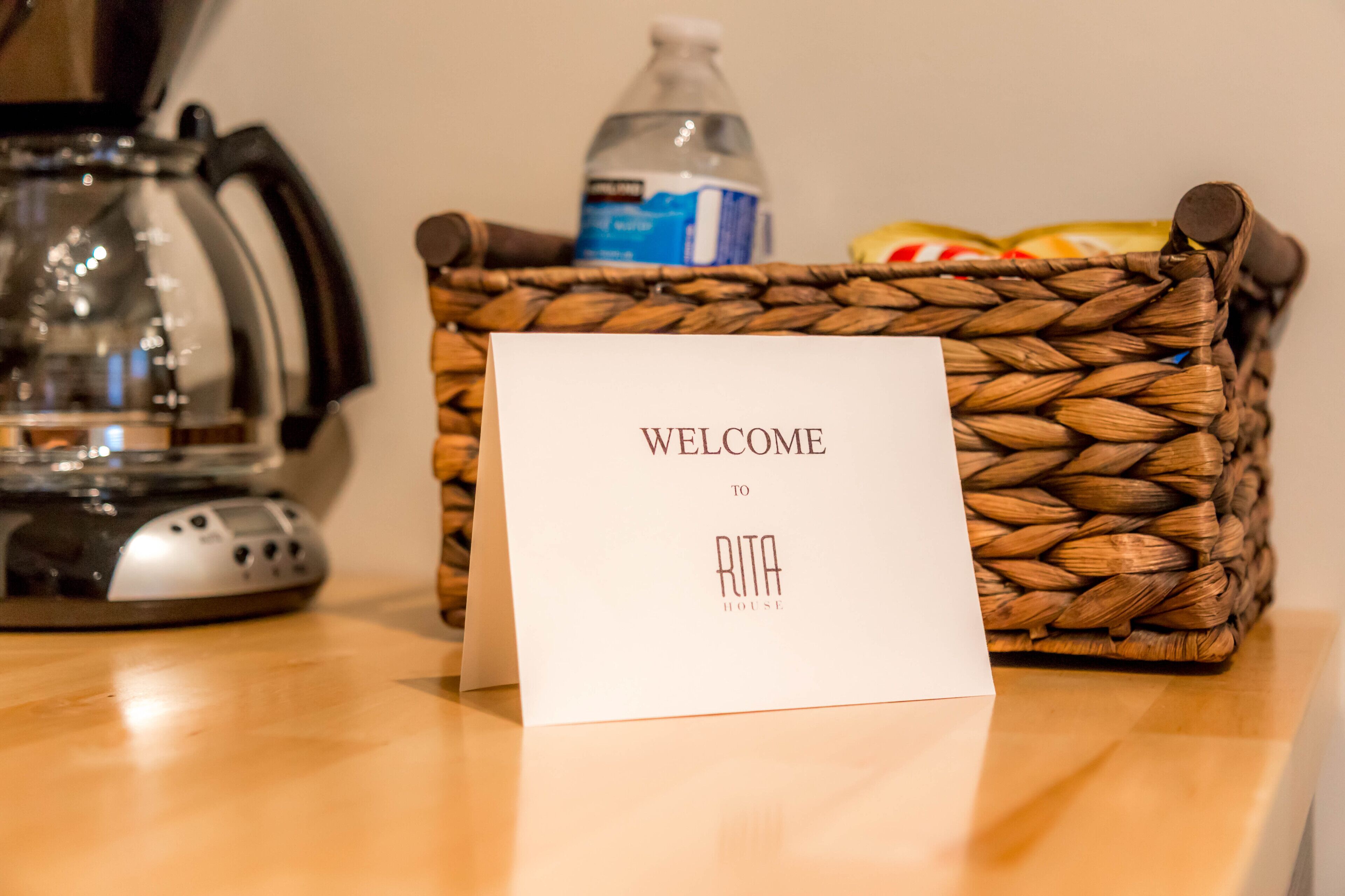 A welcome sign in a woven basket on a table with a kettle and a bottle in the background.