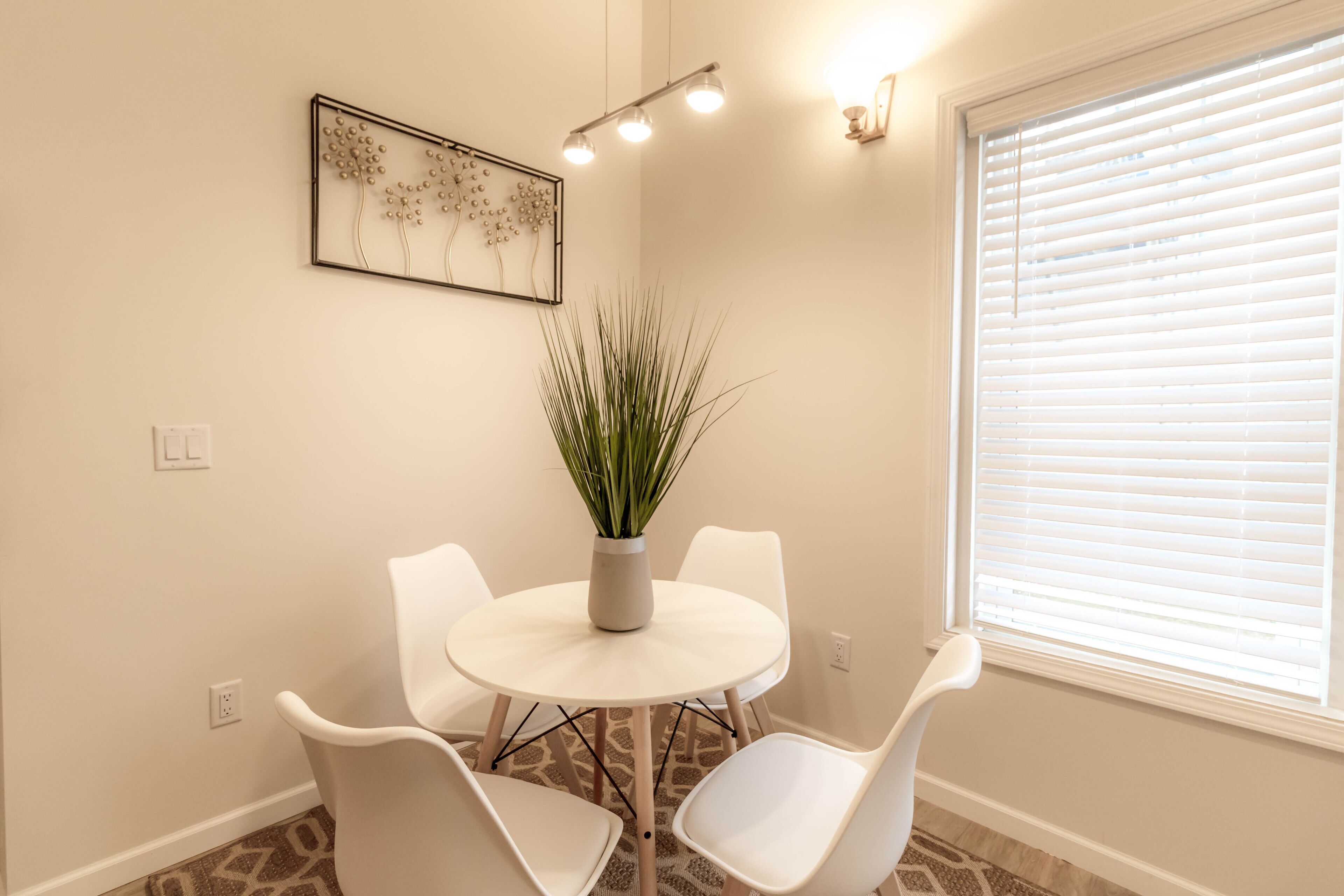 A dining area with a round table, four white chairs, a potted plant, a wall-mounted light fixture, and a window with blinds.