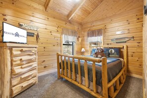 Interno di una baita in legno con un letto, una cassettiera in legno e un cartello che dice 'Qui vive un pescatore con il suo più grande trofeo'.